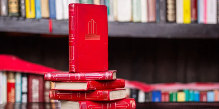 Indian-American Novels 2025: Three Remarkable Books Beyond The Migration Cliché 1 A stack of red hardcover books on a dark wooden shelf, representing Indian-American novels 2025