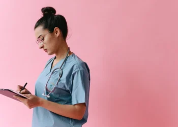 A nurse in scrubs with a stethoscope writing on a clipboard, representing the credential review an Indian nurse completes for Canada registration
